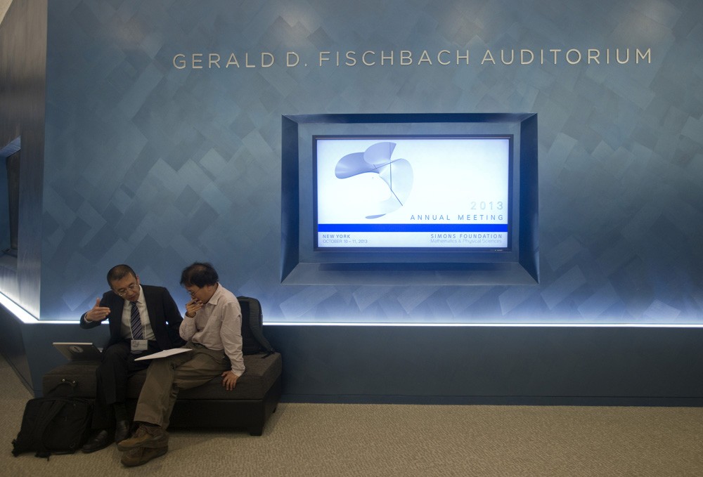 Two people sitting by a wall with 3D lettering that says Gerald D. Fischbach Auditorium