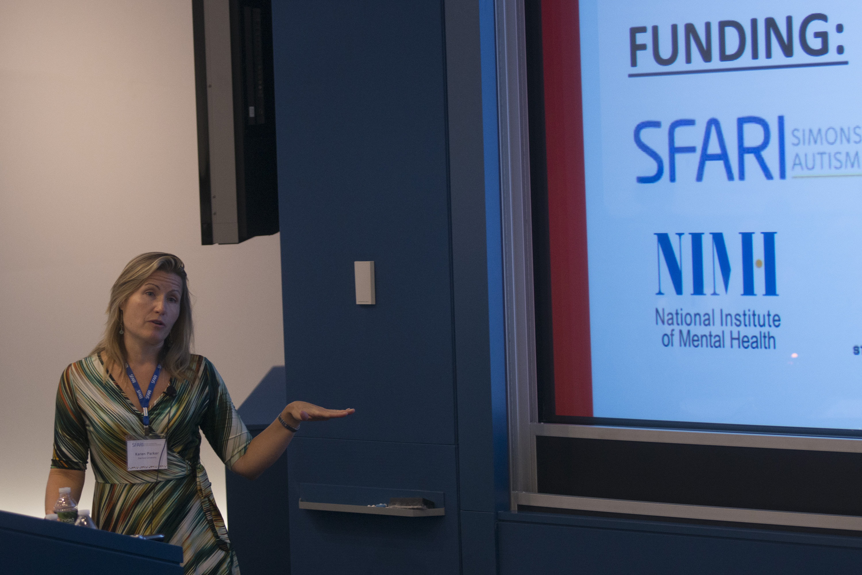Woman behind a lectern, presenting a slideshow that has SFARI and NIMH logos