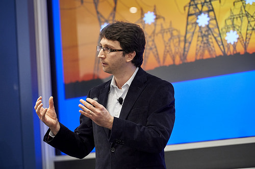 Man gesticulating as he talks on a stage with an image of power lines behind him