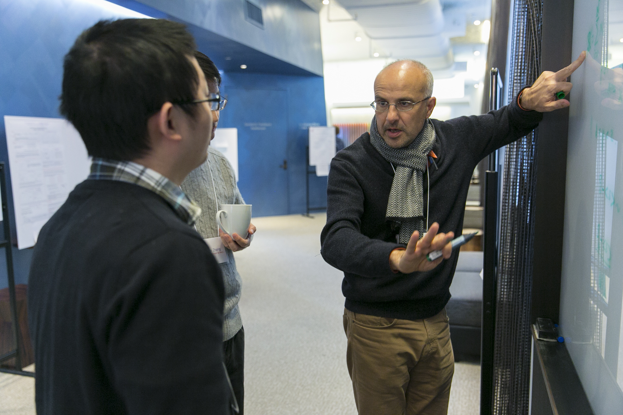 Two people listening to a man pointing to green writing on a whiteboard