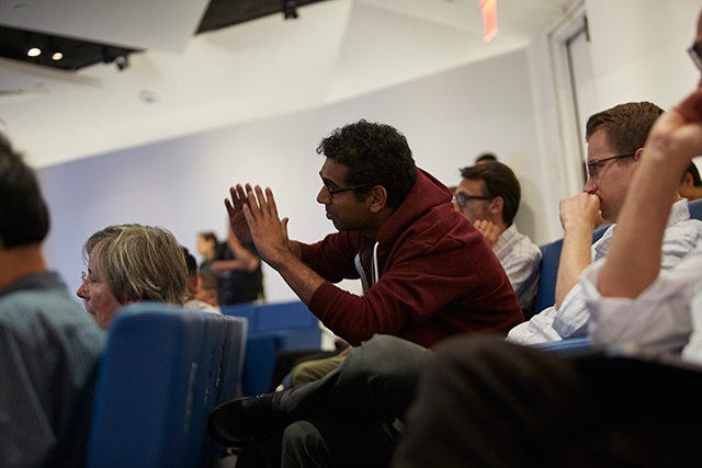 Man sitting in lecture hall seat and leaning forward while gesticulating with his hand