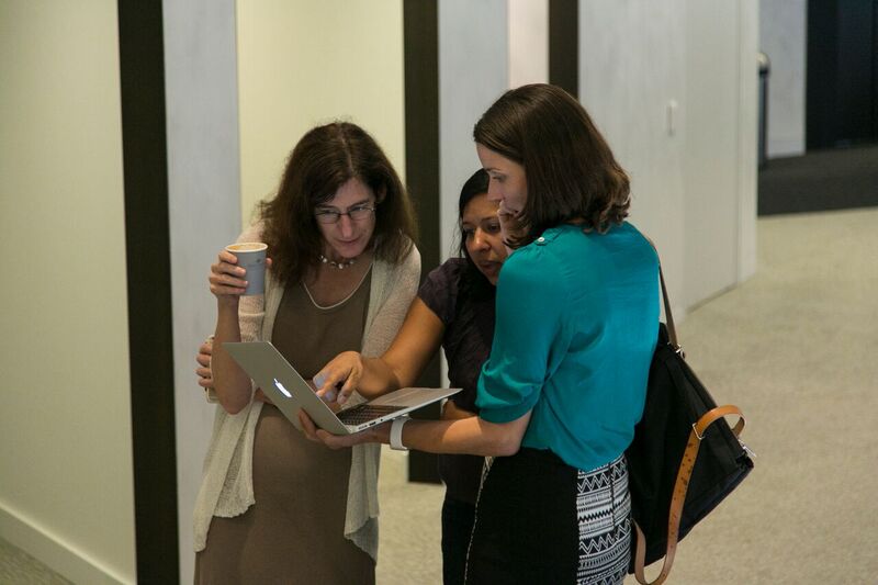 Three women standing and looking at a laptop that a woman on the right is holding