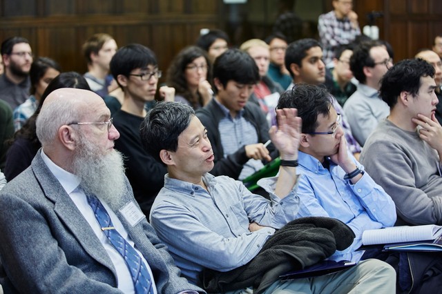 Lecture hall full of people sitting and looking forward as one man talks and gesticulates with his left hand