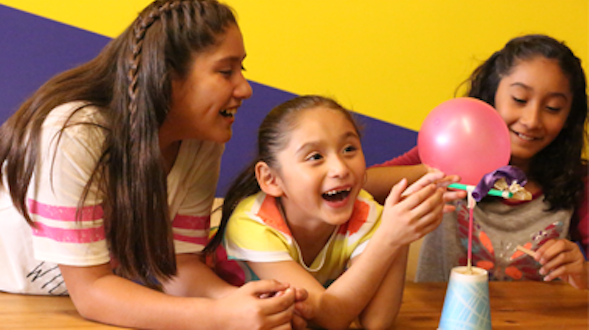 Three girls laughing at a table as a balloon connected to a cup sits in front of them