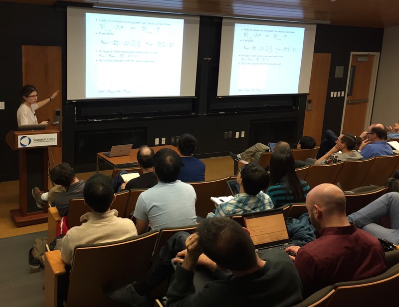 Woman standing behind a lectern in a full lecture hall as she points to a projected slideshow with math on it