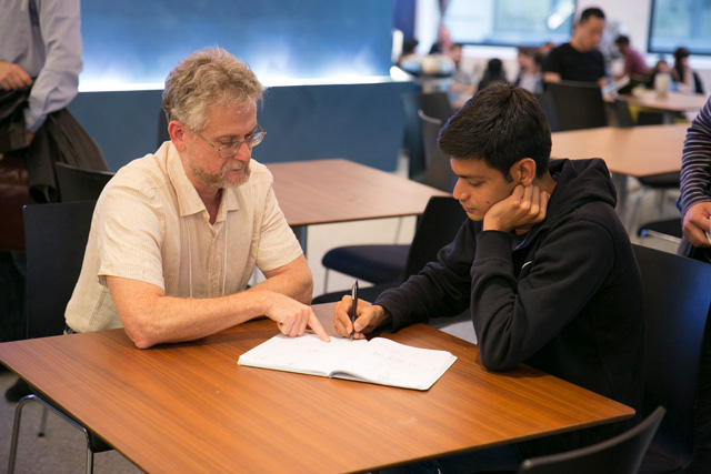 Two people sitting at a table together. One of them is pointing to the notebook the other is writing in
