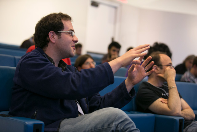 A man sitting in a lecture hall while talking with both hands outstretched