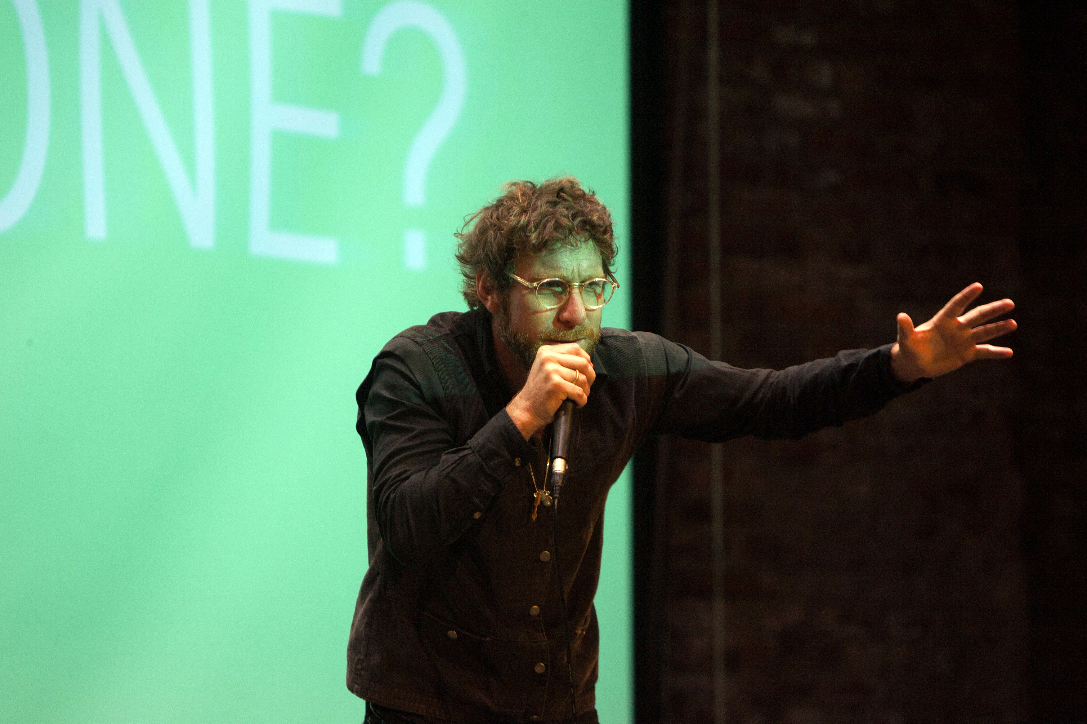 Man standing on a stage and leaning forward with an arm outstretched while the other holds a microphone to his mouth
