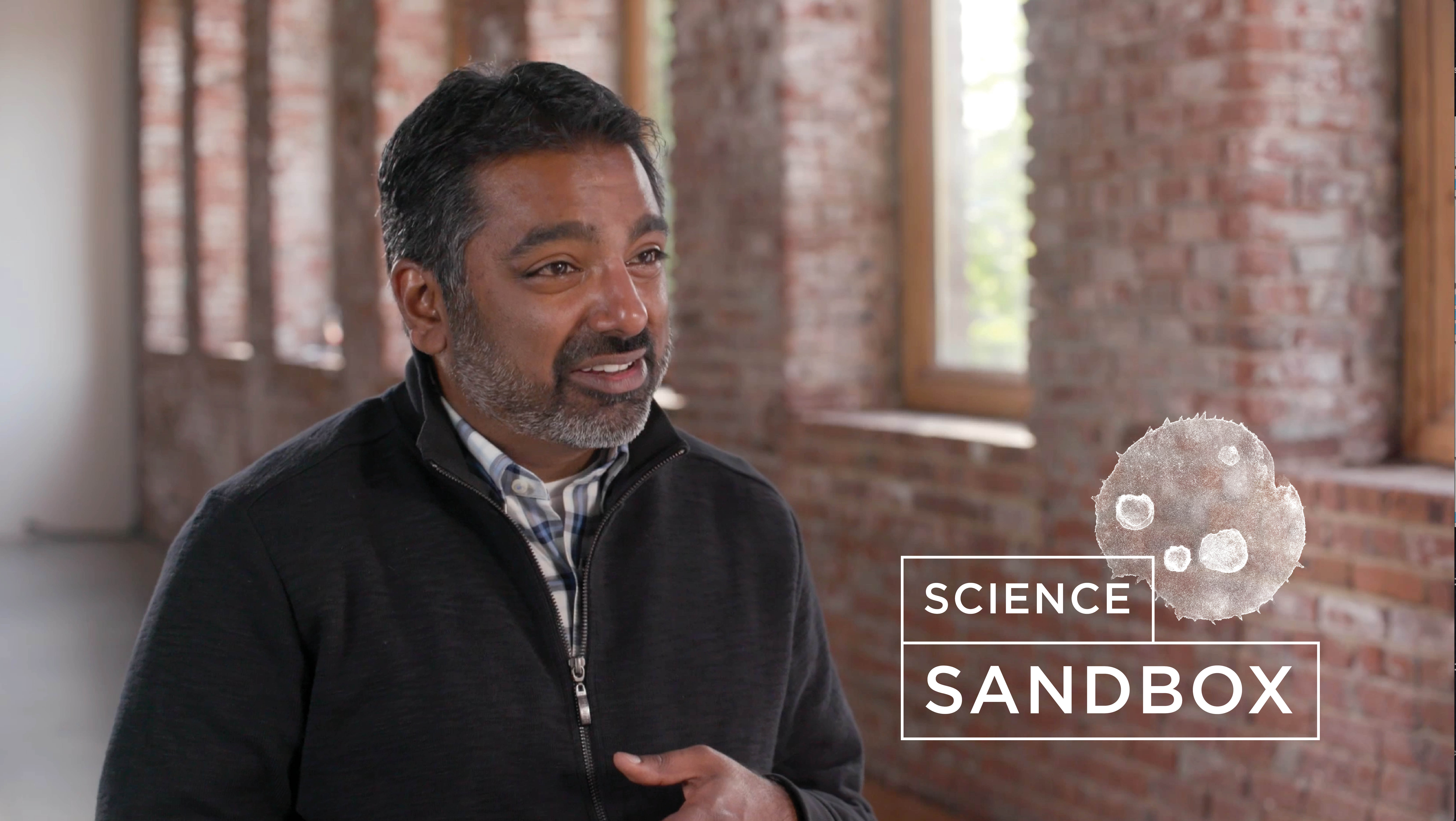 Kishore Hari talking in an empty hallway with the Science Sandbox logo pasted next to her