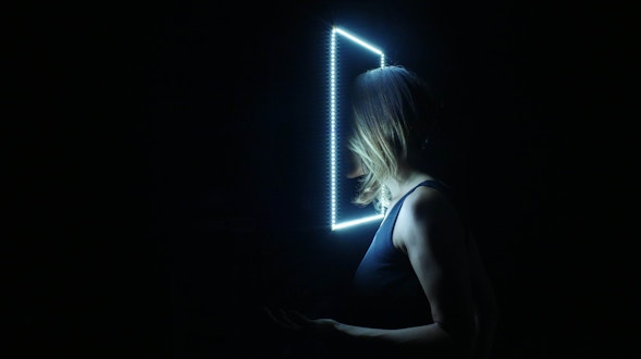 All black image with a glowing rectangle and woman putting her head through it