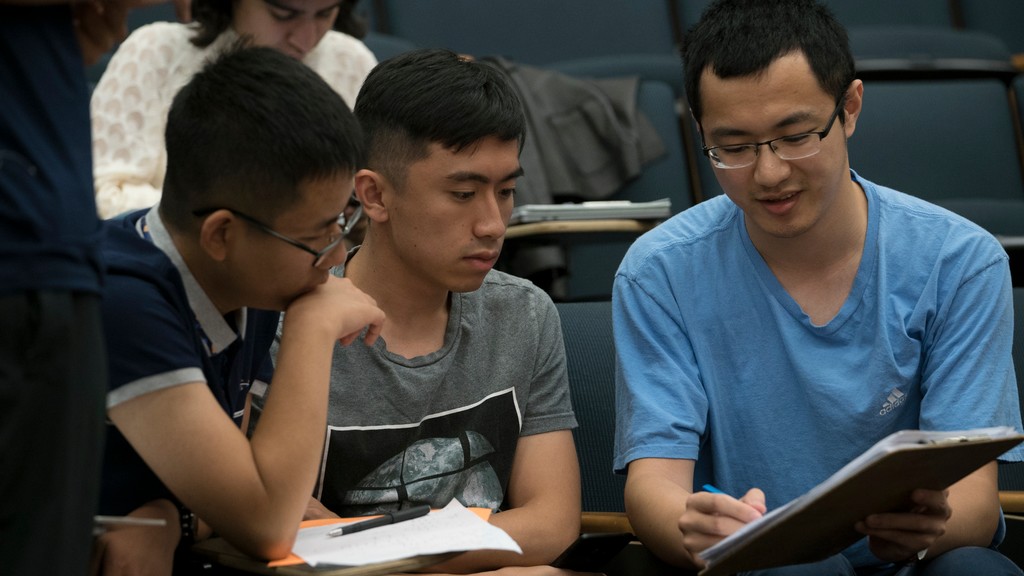 Three men sitting together in a lecture hall, looking at a notebook that the person on the right is writing in