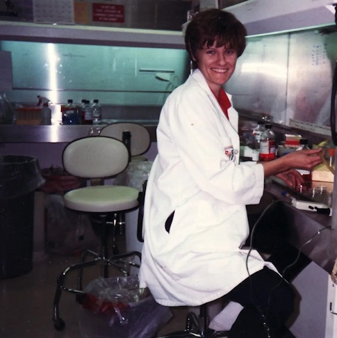 A photo of Katalin Karikó in her lab circa. 1989. She is a young woman with short brown hair, a red top, a white lab coat, and black pants. She smiles while looking towards the camera and handling a brownish tube-like object with her right hand at her desk. The lab itself is a room with a dark-colored floor, multiple waste bins, at least three beige wheeled stools, including one of which that Karikó is sitting on, and a steel countertop lining the reflective walls of the lab. The section of the countertop that Karikó is working at has multiple bottles, cartons, and a white device connected to a wire that holds several small glass containers. Another section of the countertop, located towards the right of the photograph, has multiple clear bottles and containers, including one with a spray nozzle. The walls are reflective, but opaque--the section that Karikó is close to has illegible writing on it, while the section on the right is mostly empty save for a rectangular strip stuck onto it and holding a scrap of paper. Above the section of the wall on the strip of the photo is a white poster resembling a counter and a red sign with illegible white text on it.