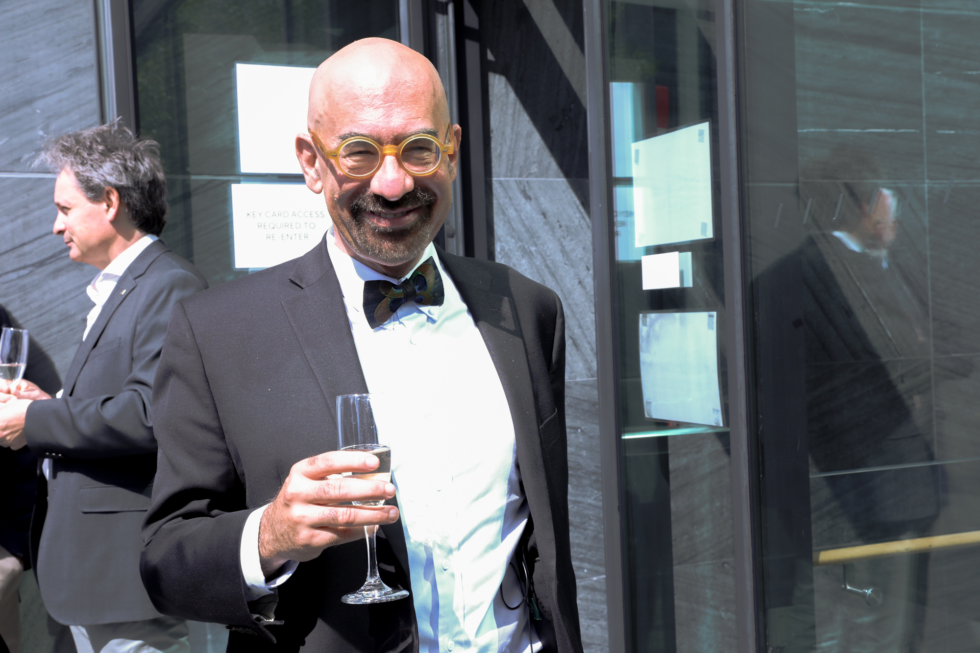 A photograph of David Spergel, a bald bearded man with yellow circular glasses, in a black suit with a white shirt and black bowtie. He is situated in an outdoor setting where there are people similarly dressed to him standing in the background. He is smiling at the camera and is holding a flute of champagne.