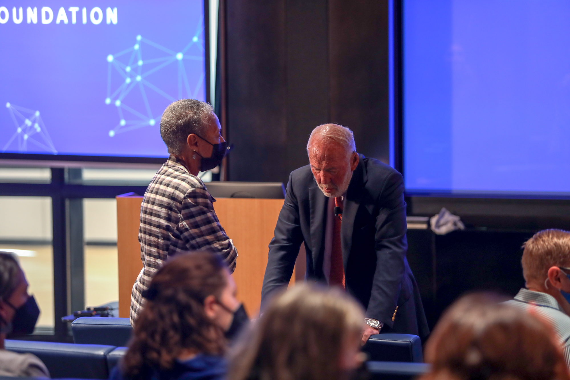 A photograph of two people, a woman wearing a mask and a man without a mask and propping himself up with a chair, in an auditorium.