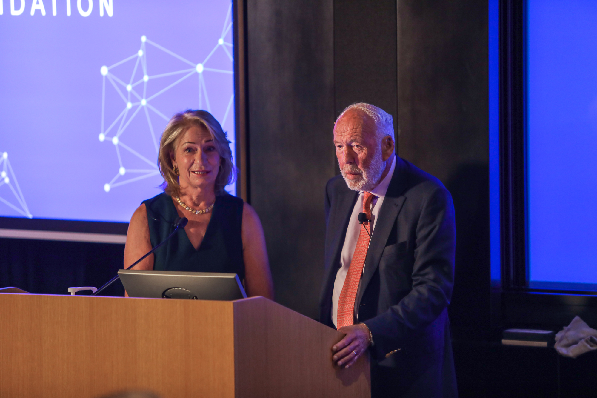 A photograph of two people standing beside a wooden podium and a screen with a slide projected onto it.