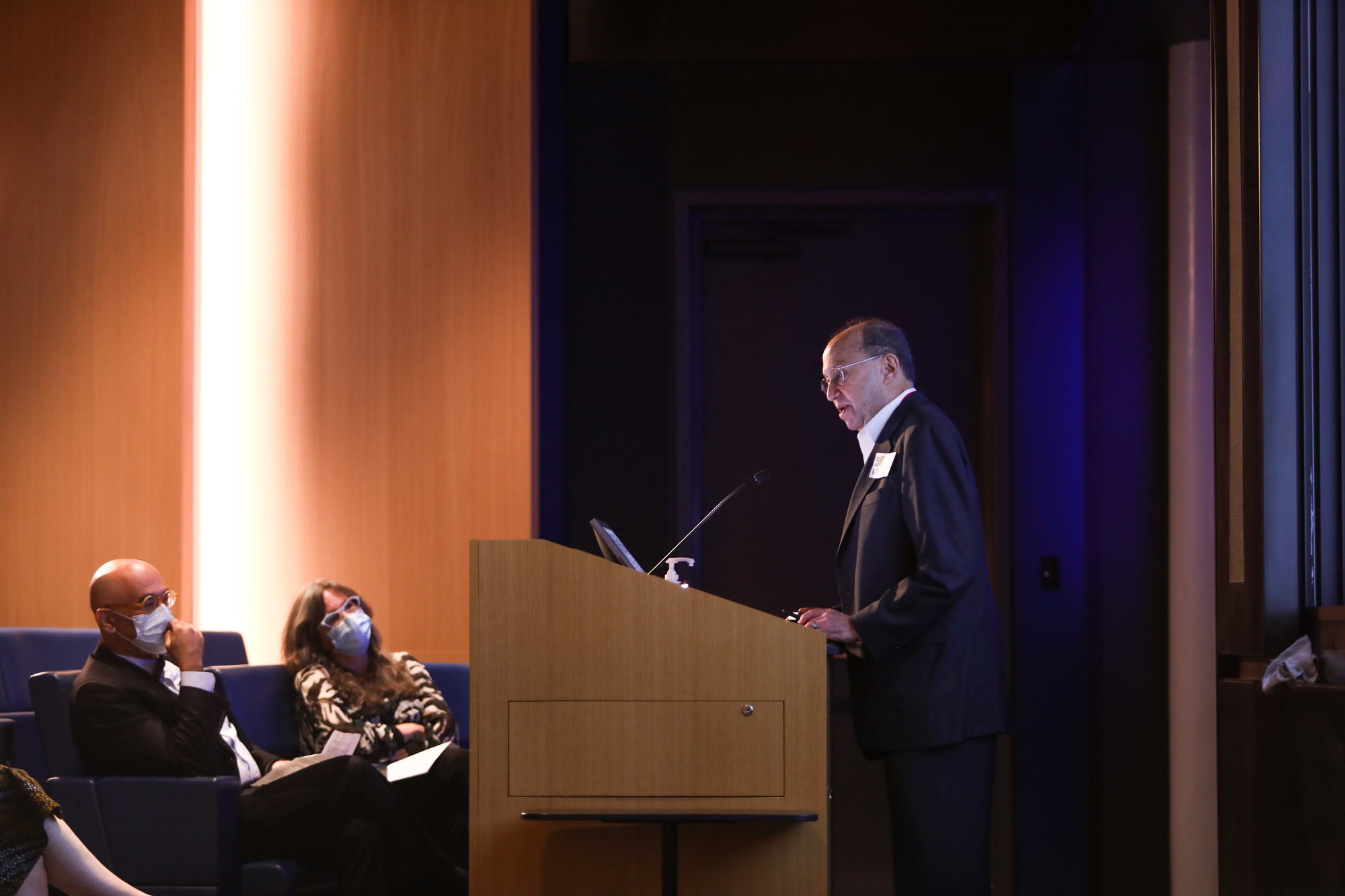 A photograph of a man speaking at a wooden podium as two people wearing masks sit in auditorium seats.