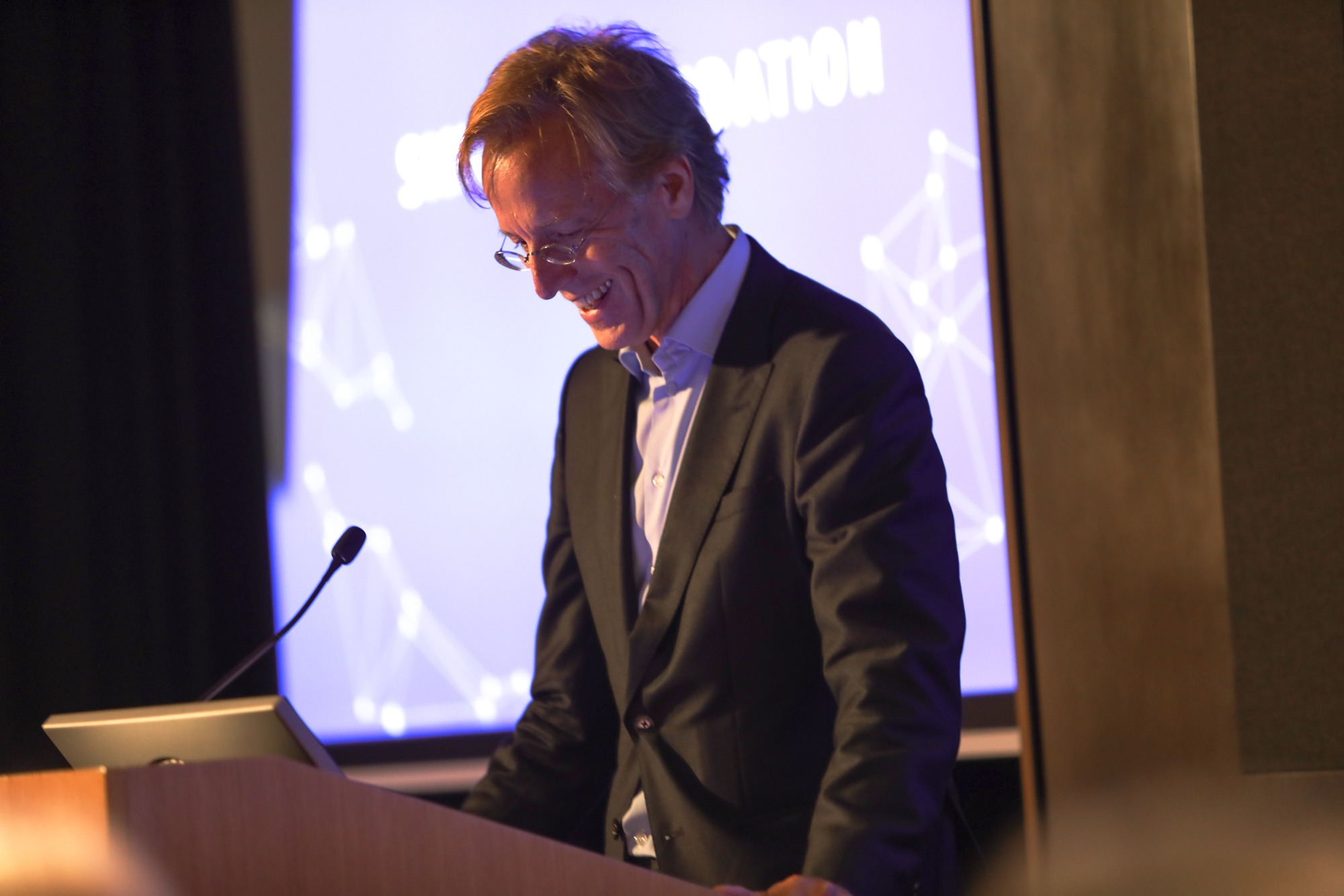 A photograph of a person speaking at a wooden podium and standing beside a screen with a slide projected onto it.