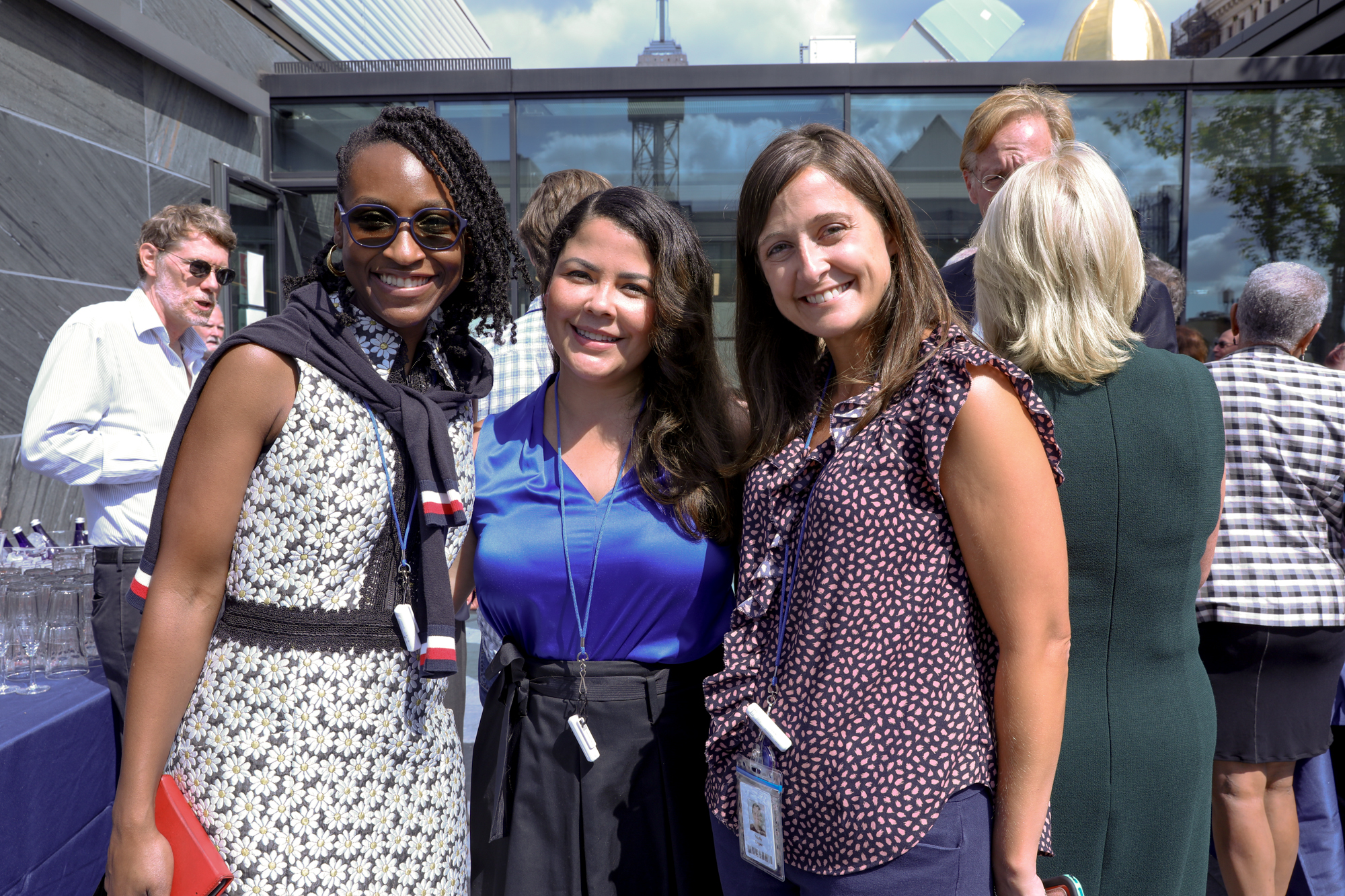 A photograph of three women smiling towards a camera as they stand in an outdoor space.