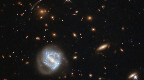 乍一看，这张照片被画面左下角旋转螺旋的明亮光芒所主导。然而，这个星系远非这里最有趣的景象——它的后面是一个星系团。星系在空间中不是随机分布的;它们在地心引力的强力作用下聚集在一起，形成群体和集群。银河系是本星系群的一员，本星系群是室女座星系团的一部分，室女座星系团又是拥有10万个星系的拉尼亚克亚超星系团的一部分。这张图片中看到的星系团被称为SDSS J0333+0651。像这样的星系团可以帮助天文学家了解遥远的——因此也是早期的——宇宙。SDSS J0333+0651的成像是对遥远星系恒星形成研究的一部分。恒星形成区域通常不是很大，最多延伸几百光年，所以望远镜很难在远处分辨它们。即使使用它最灵敏、分辨率最高的相机，哈勃望远镜也无法探测到非常遥远的恒星形成区域，因此天文学家使用了一种宇宙技巧:他们转而寻找星系团，星系团的引力影响如此之大，以至于它们扭曲了周围的时空。 This distortion acts like a lens, magnifying the light of galaxies sitting far behind the cluster and producing elongated arcs like the one seen to the left of centre in this image.