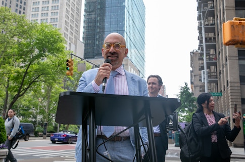 David Spergel, President of the Simons Foundation, dressed in a light blue suit, yellow circle-rimmed glasses and a light pink shirt, holding a mic and standing at a podium