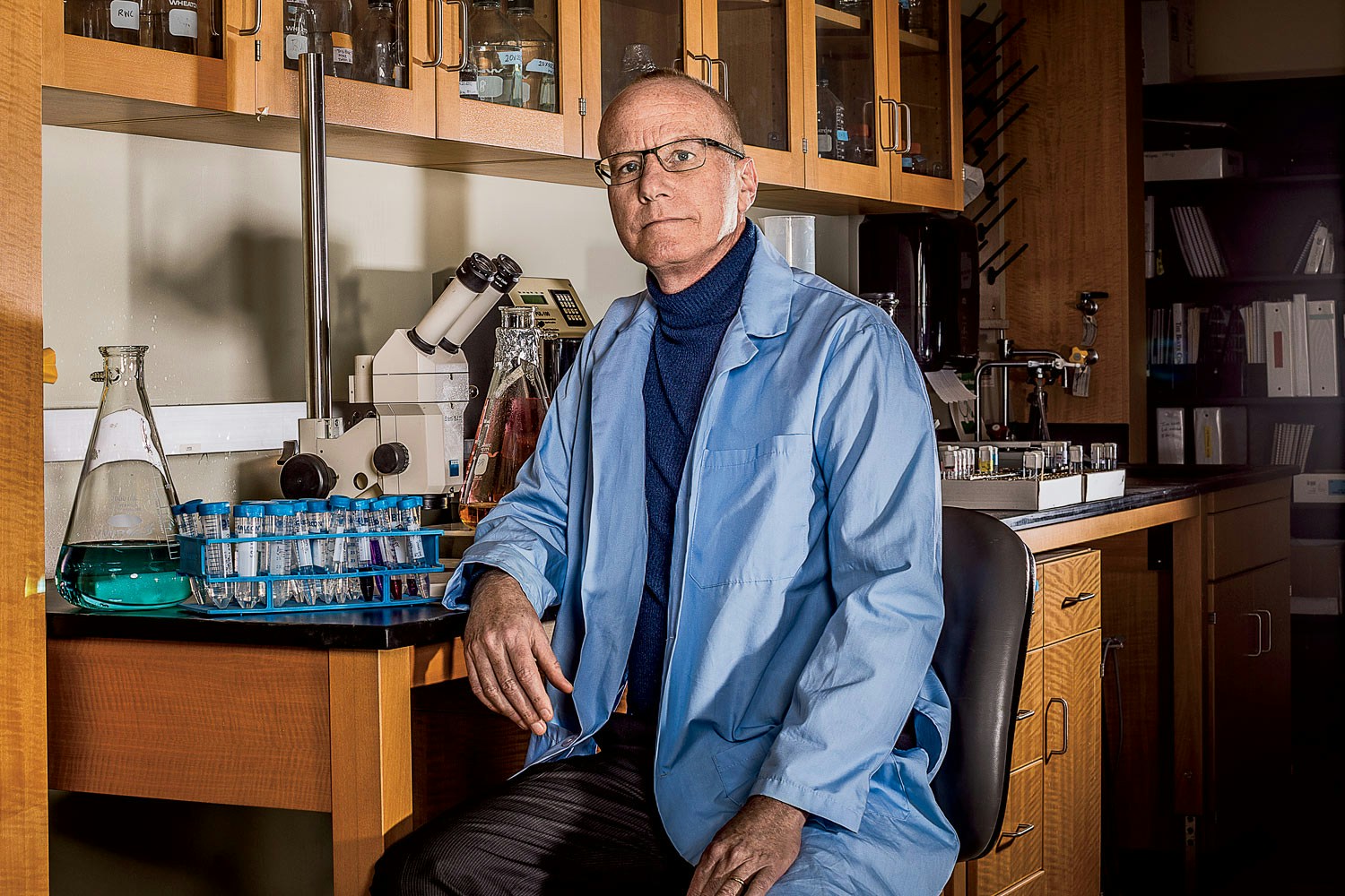 Man wearing rectangular eyeglasses and a jacket sitting on a stool next to a table with a microscope and test tubes