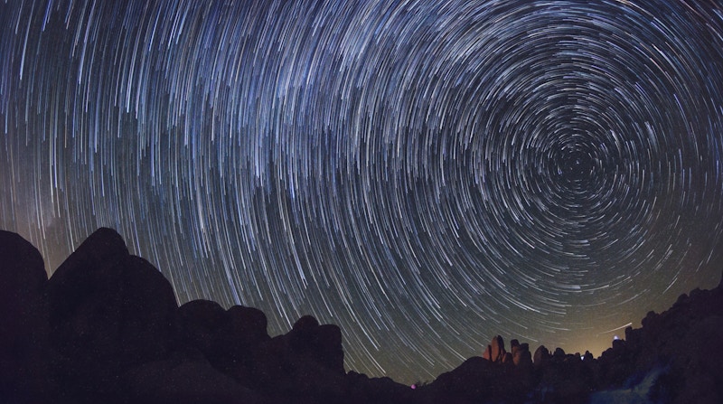 Slow exposure of night sky with stars swirling over a darkened landscape.