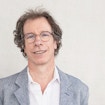 Portrait of Stéphane Mallat, winner of the 2025 CNRS Gold Medal, wearing a grey suit and leaning against a wall.
