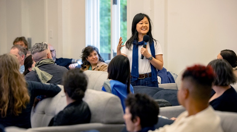 A woman speaks into a microphone to a seated audience, who turn to look at her.