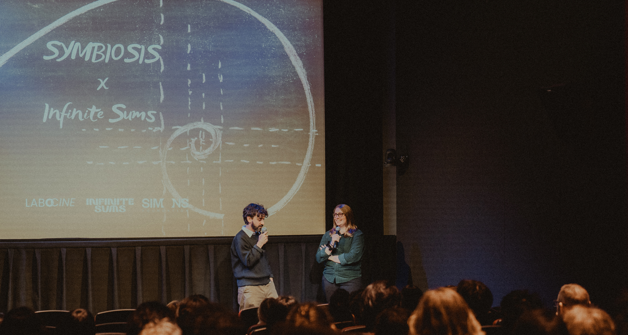 A photo of Sonya Hansen and Diego Murillo at the Symbiosis film screening, standing in front of a screen that says Symbiosis x Infinite Sums.