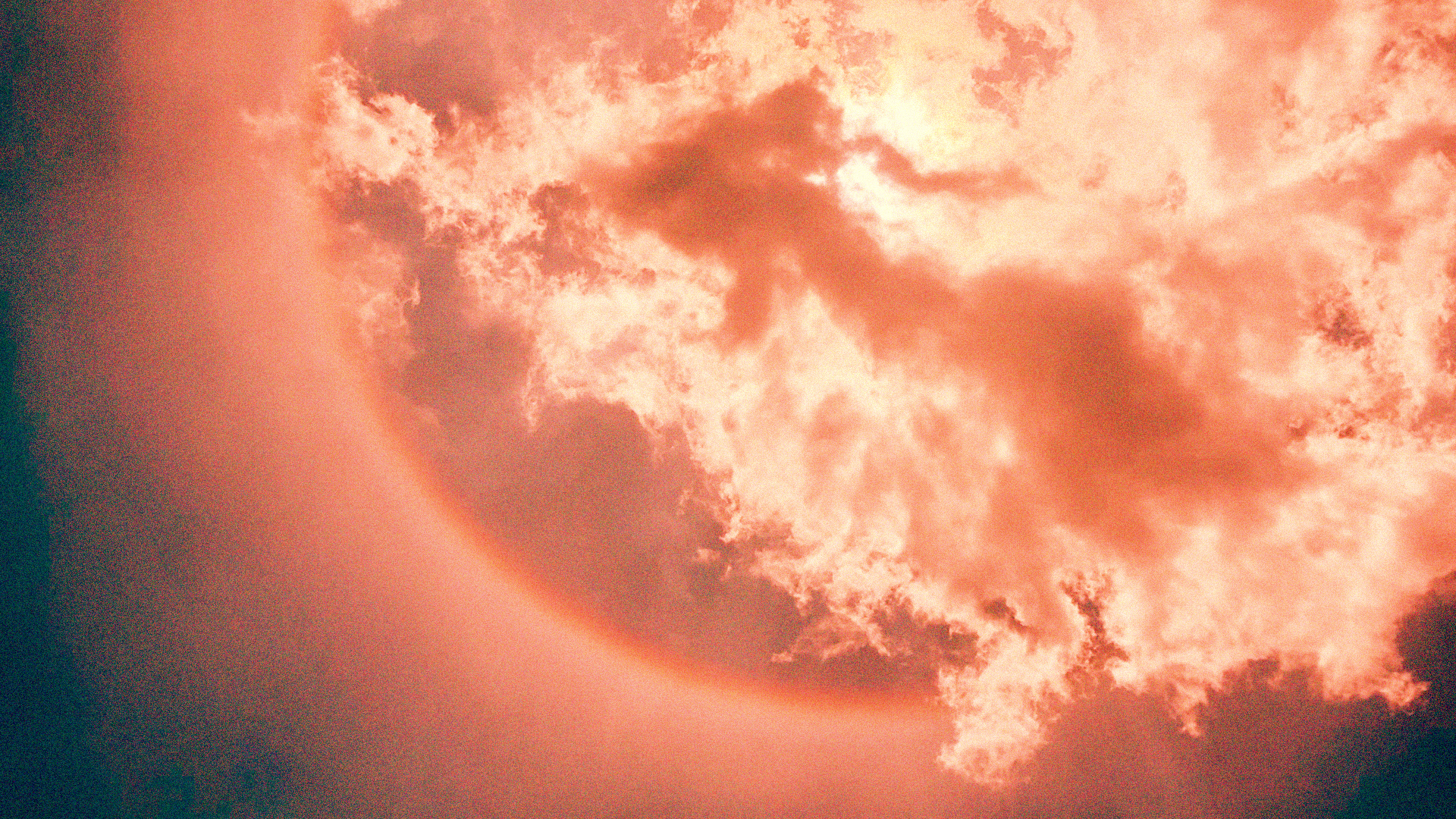 Still image from Absolute Zero: A full moon partially obscured by dramatic, billowing clouds, bathed in deep coral and orange hues, creating an ethereal and moody atmosphere.