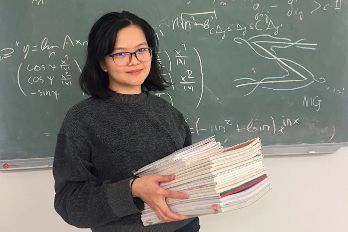 A portrait of Gervy Marie Angeles holding a pile of books, standing in front of a blackboard.