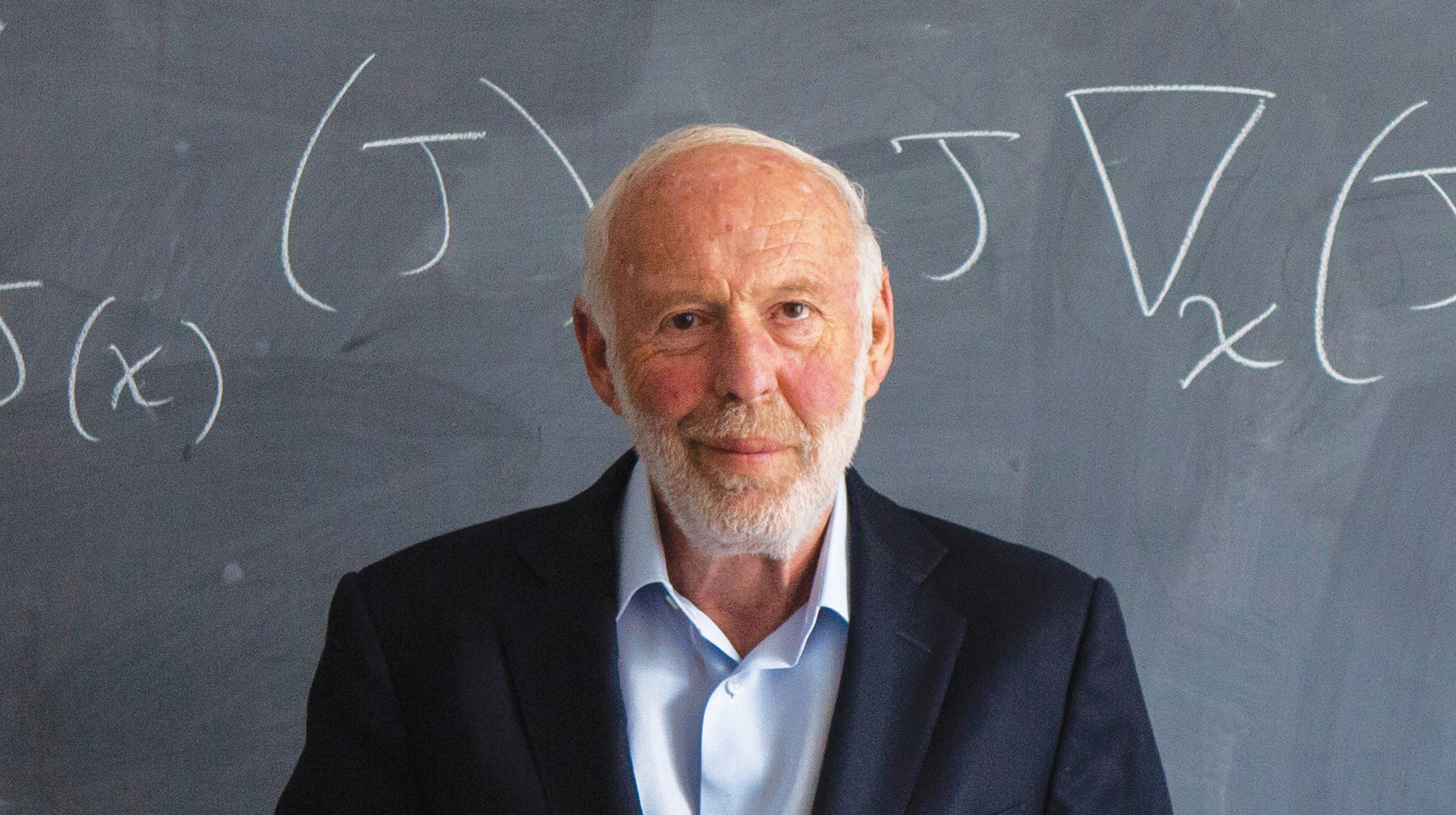 Photo of Jim Simons in front of a chalk board that shows an equation.