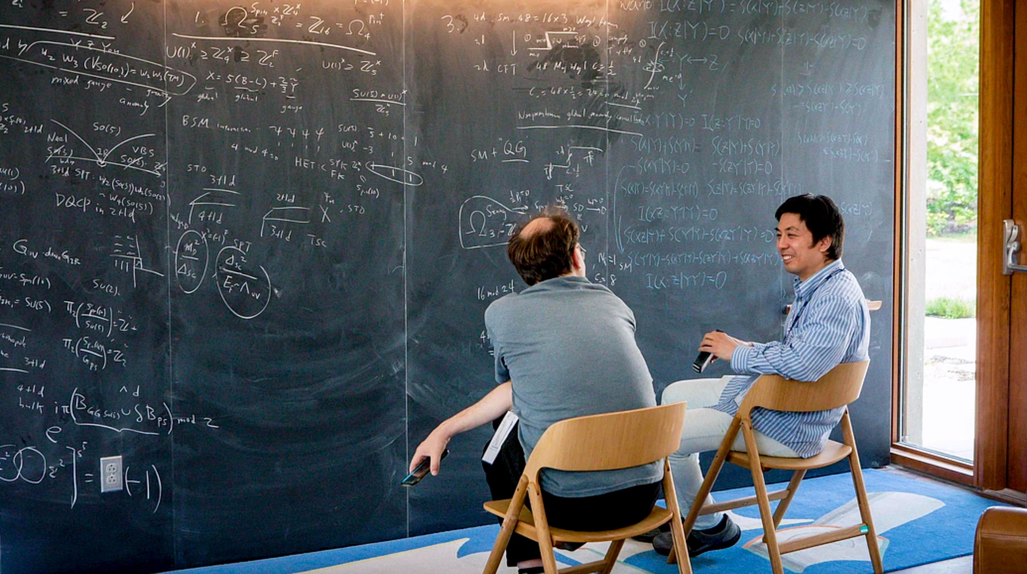 A photo of two people sitting and discussing in front of a chalkboard.