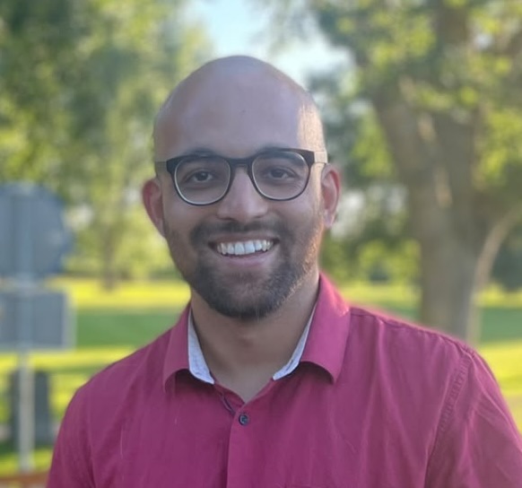 A smiling person with glasses and a beard, wearing a red collared shirt, stands outdoors with green trees and sunlight in the background.