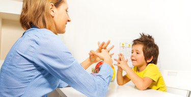 Boy exercises by putting hands and fingers together with therapist showing it improving motor skills sitting opposite at the table indoors